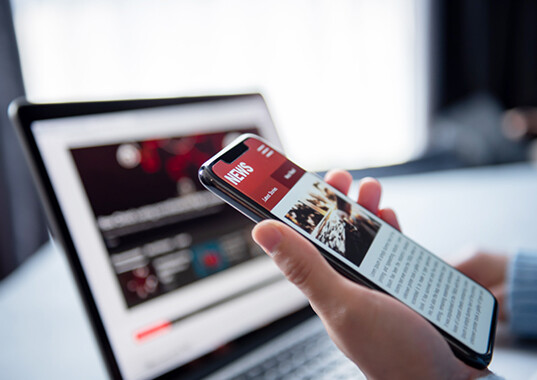 Person reading news on a smartphone beside a laptop.