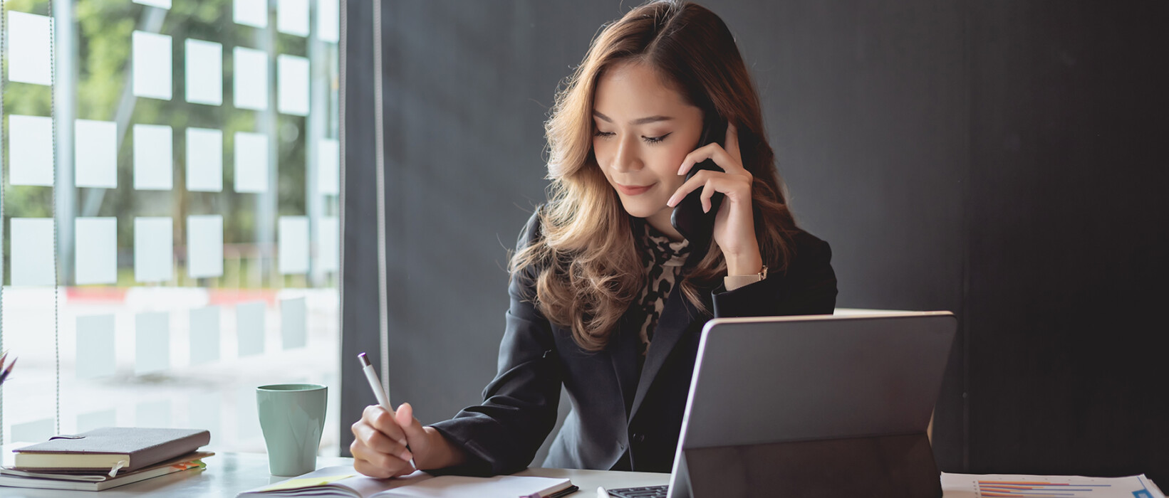 Business professional on the phone while writing notes at a desk.